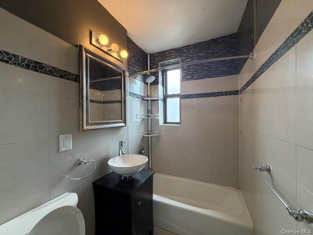 Bathroom featuring a white bathtub, white wall tiles with decorative blue accents, a pedestal sink with a dark cabinet, and a window for natural light
